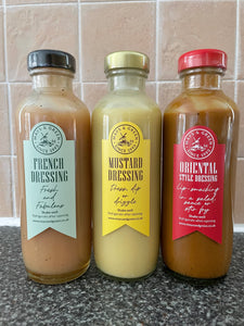 Three glass bottles of salad dressing; Mustard with a light yellow label, Oriental with a light red label and French with a light green label.
